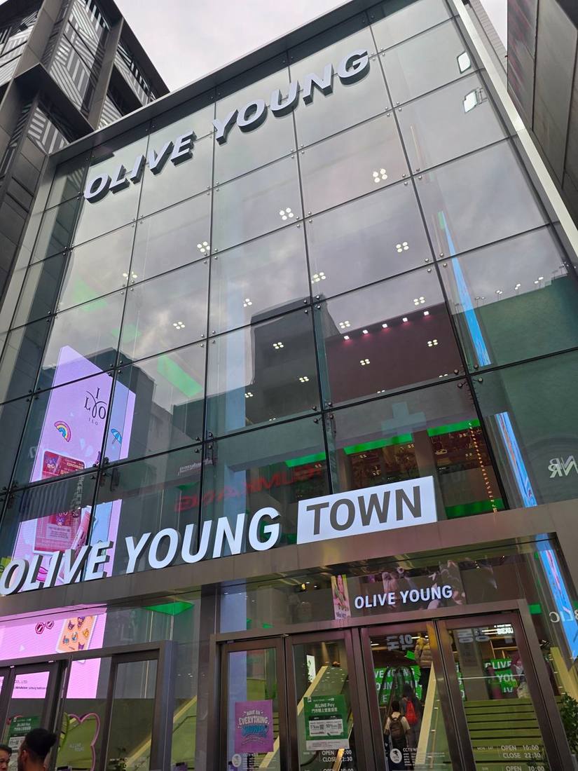 4개 층 높이 통유리 커튼월에 ‘OLIVE YOUNG’ 대형 레터와 1층 ‘OLIVE YOUNG TOWN’ 사인을 단 플래그십. 유리 너머로 녹색 에스컬레이터와 다층 매장이 보이고, 왼쪽에 대형 세로 LED, 입구에 영업시간(10:00~22:30)과 LINE Pay 프로모션 안내가 있습니다.